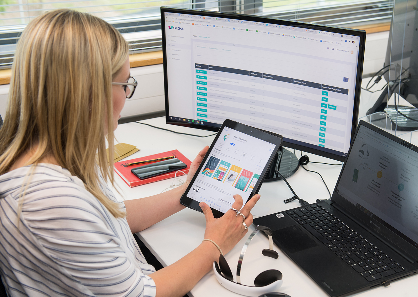 ORCHA digital health assessor holding an iPad, looking at ORCHA review engine on a laptop and computer screen.
