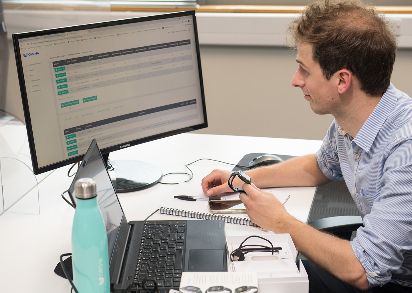 ORCHA digital health assessor viewing the Review Engine on a desktop computer, with laptop open in front of him
